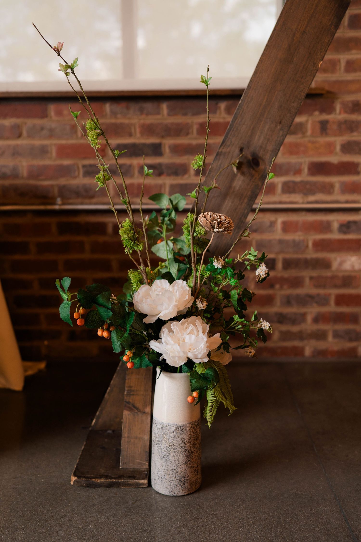 White peony floral arrangement in ceramic vase beside rustic wooden beam at brick wall venue.