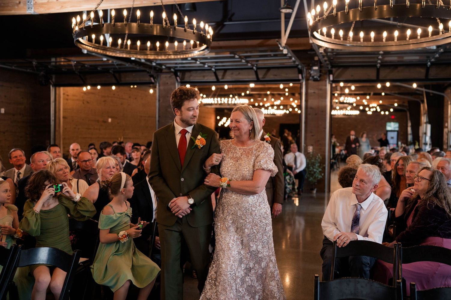 Groom escorts woman in lace dress down wedding ceremony aisle amid applauding guests.