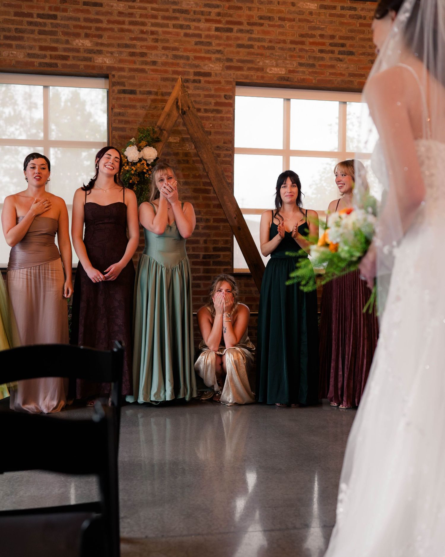 Emotional bridesmaids in earth-tone gowns reacting with tears as bride approaches in white gown.