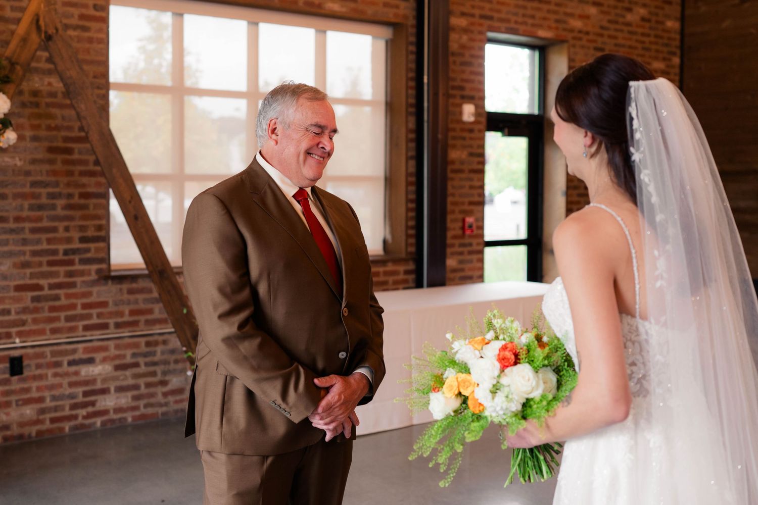 Father of bride in tan suit smiling at daughter in white wedding gown during first look.