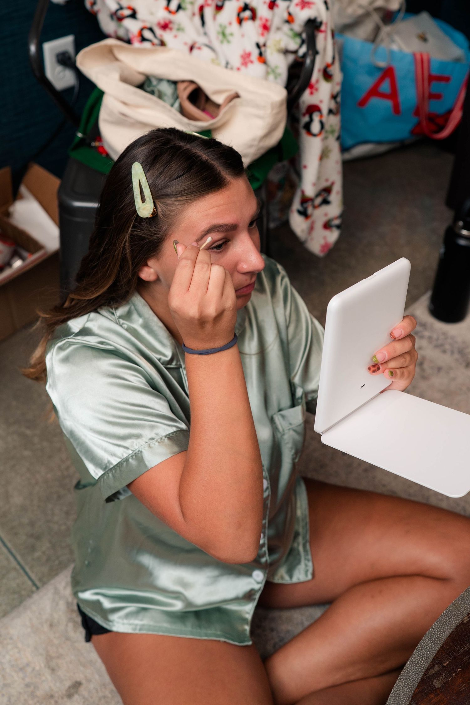 A young woman in a green satin pajama top applies makeup using a small handheld mirror.