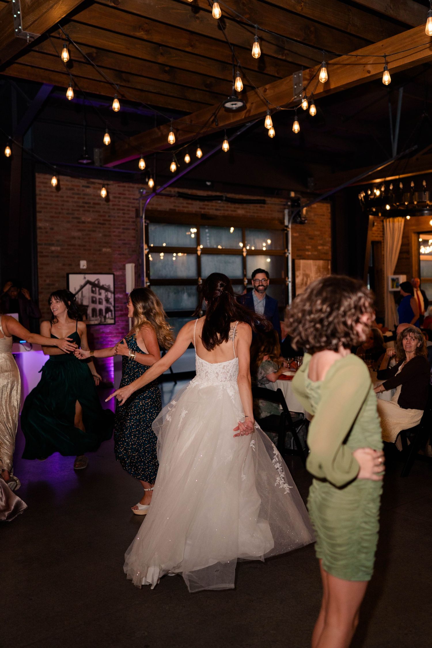 Bride in white tulle gown dancing with guests in rustic brick wedding venue with Edison bulb lights.