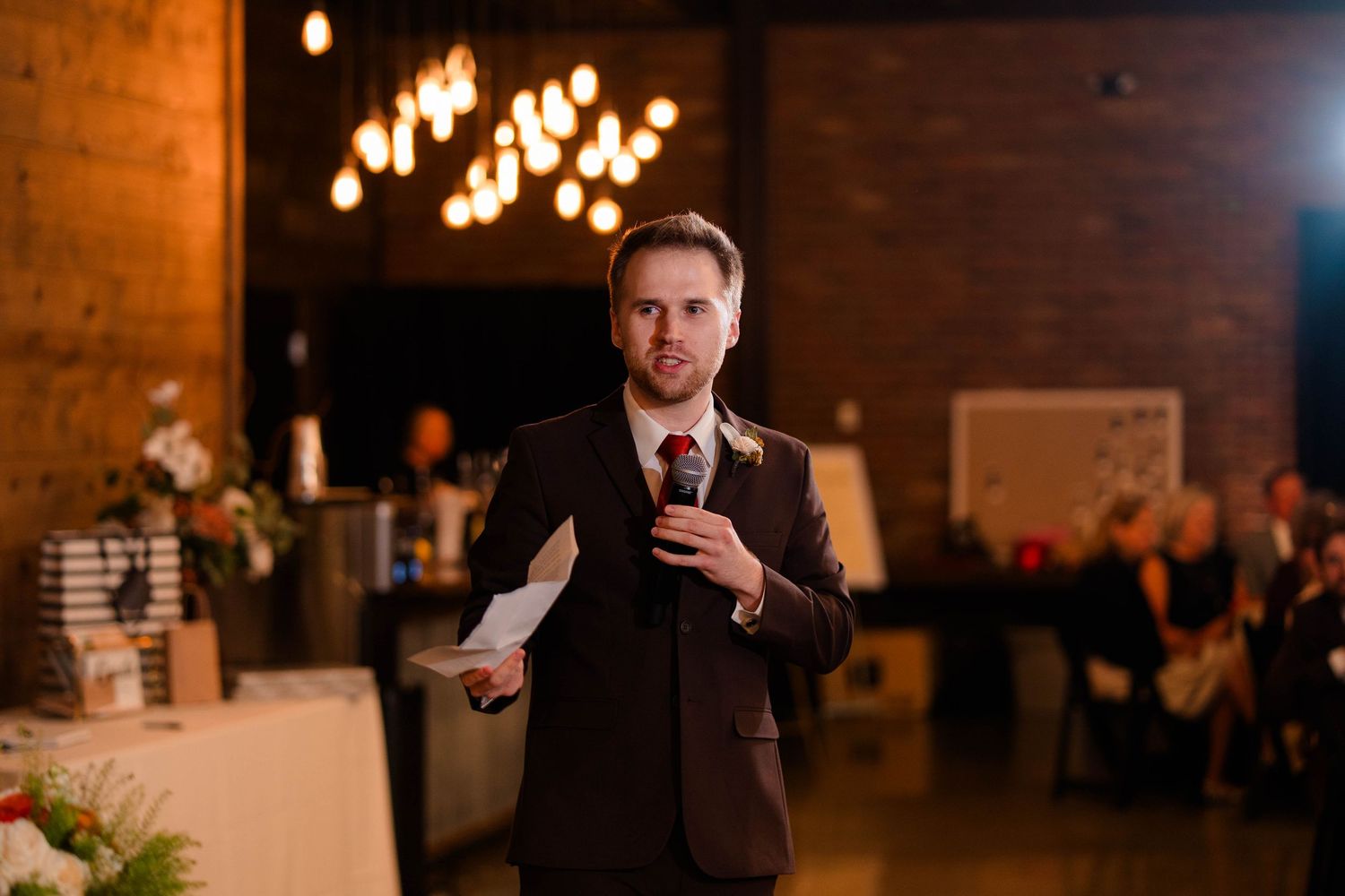 Best man delivers heartfelt wedding toast holding paper notes in decorated industrial reception venue.