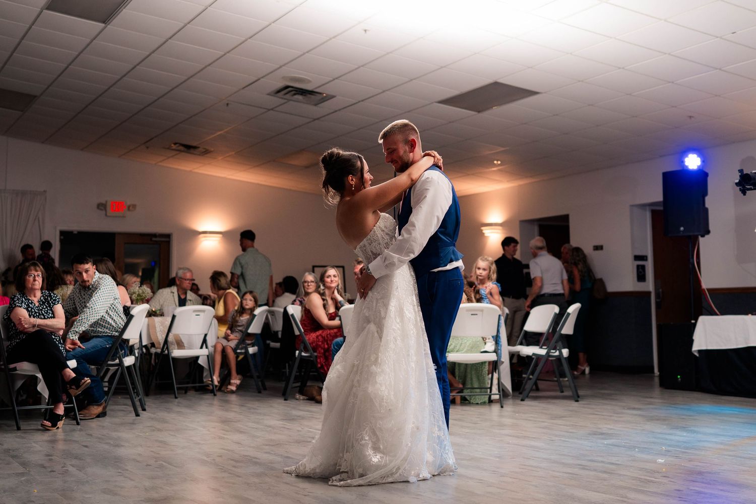 Bride and groom share their first dance at wedding reception while guests watch from white chairs.