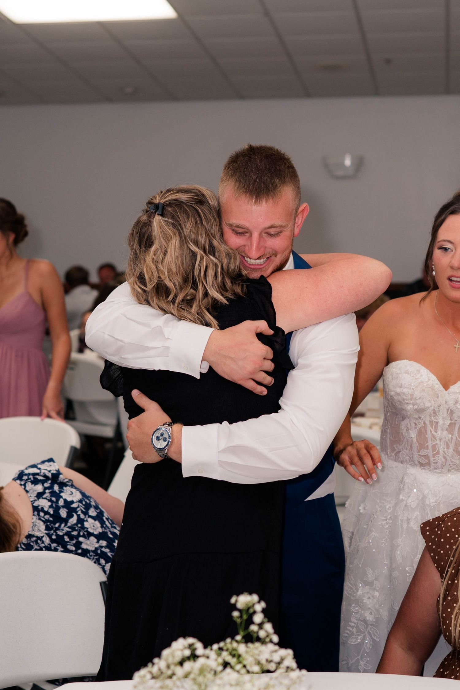 Groom in navy suit smiling while sharing a warm hug with a guest at a wedding reception.