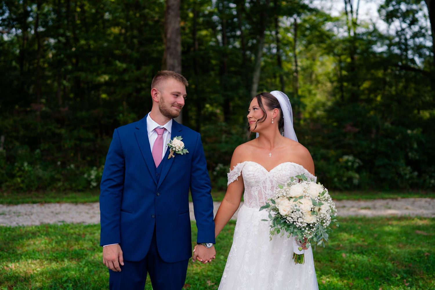 Bride and groom holding hands smiling at each other outdoors in a lush green forest setting.