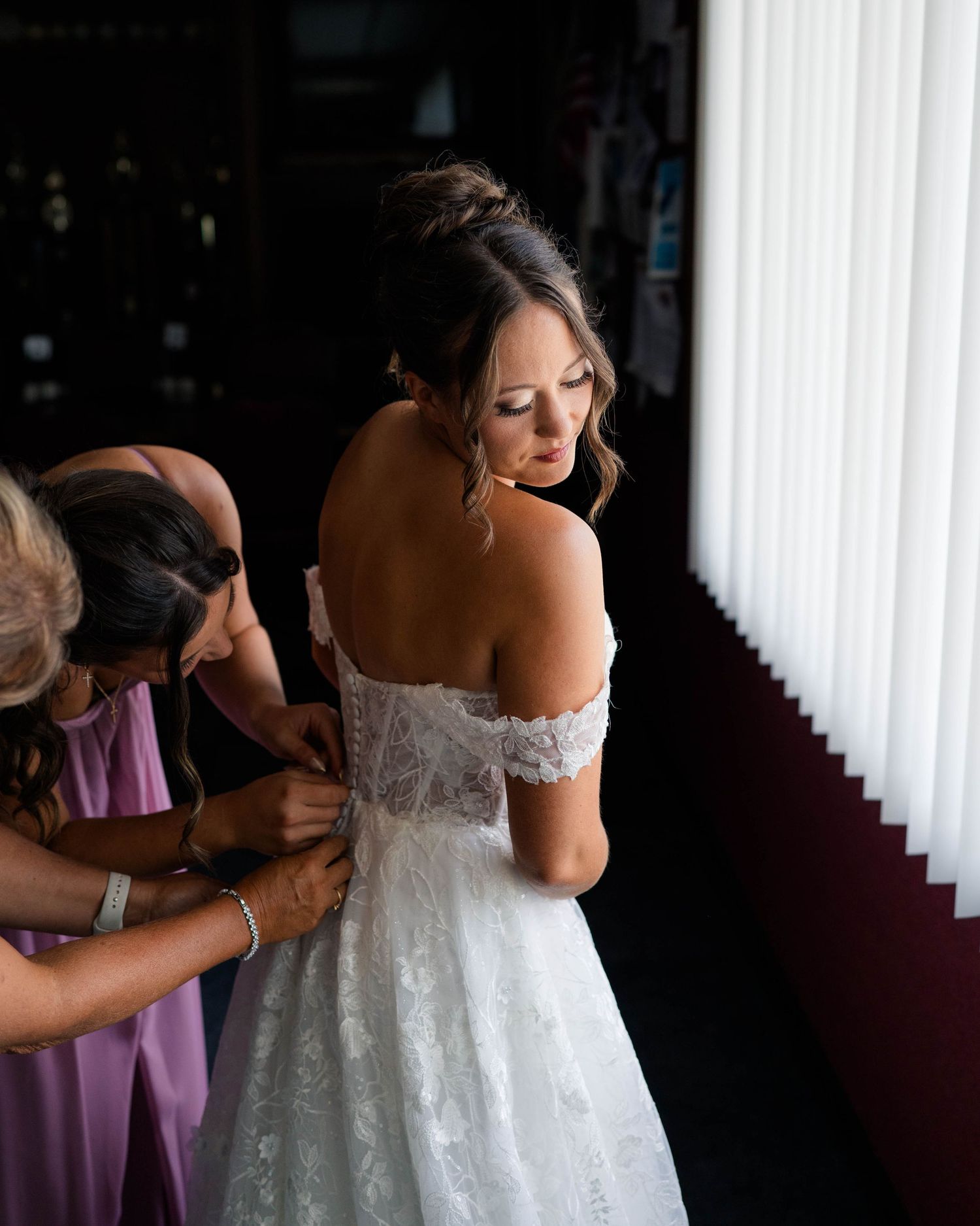 Bride in off-shoulder lace wedding gown being buttoned by bridesmaids in lavender dresses.