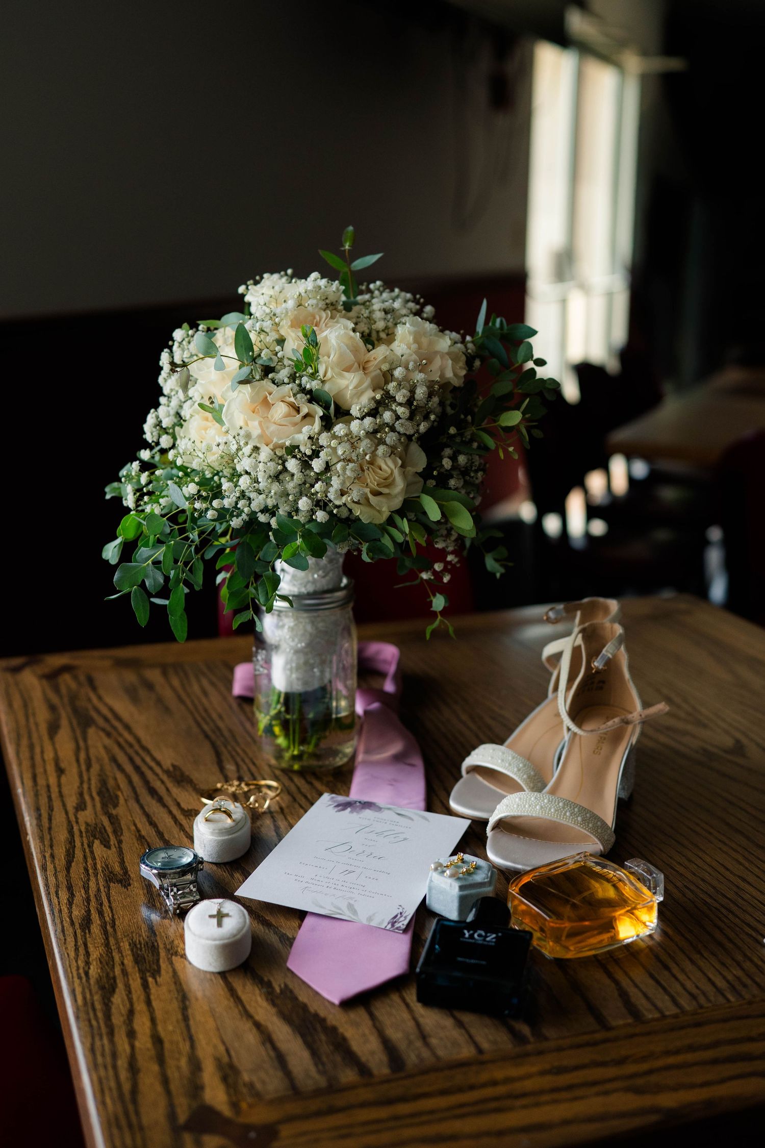 Bridal flat lay with white floral bouquet, heels, perfume, rings, and pink tie on wooden table.