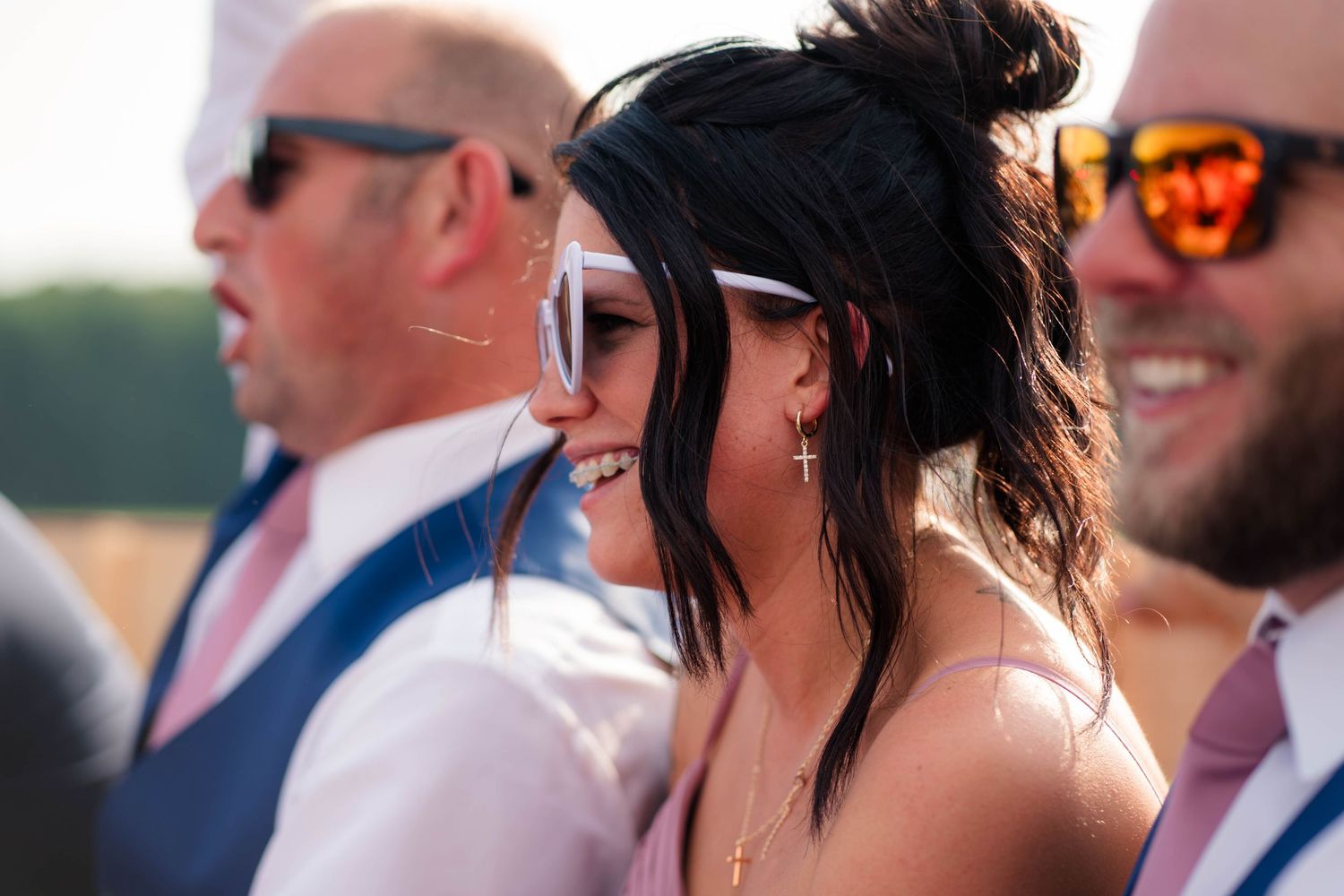 Wedding guest smiling in white heart sunglasses with cross necklace at outdoor summer wedding celebration.