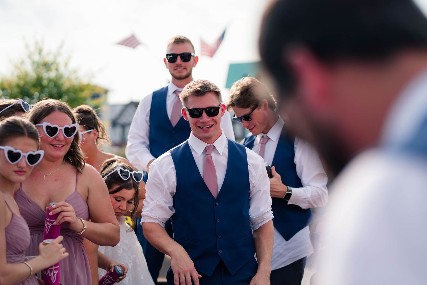 Wedding guests walking outdoors, groomsmen in navy vests with pink ties wearing sunglasses at reception.
