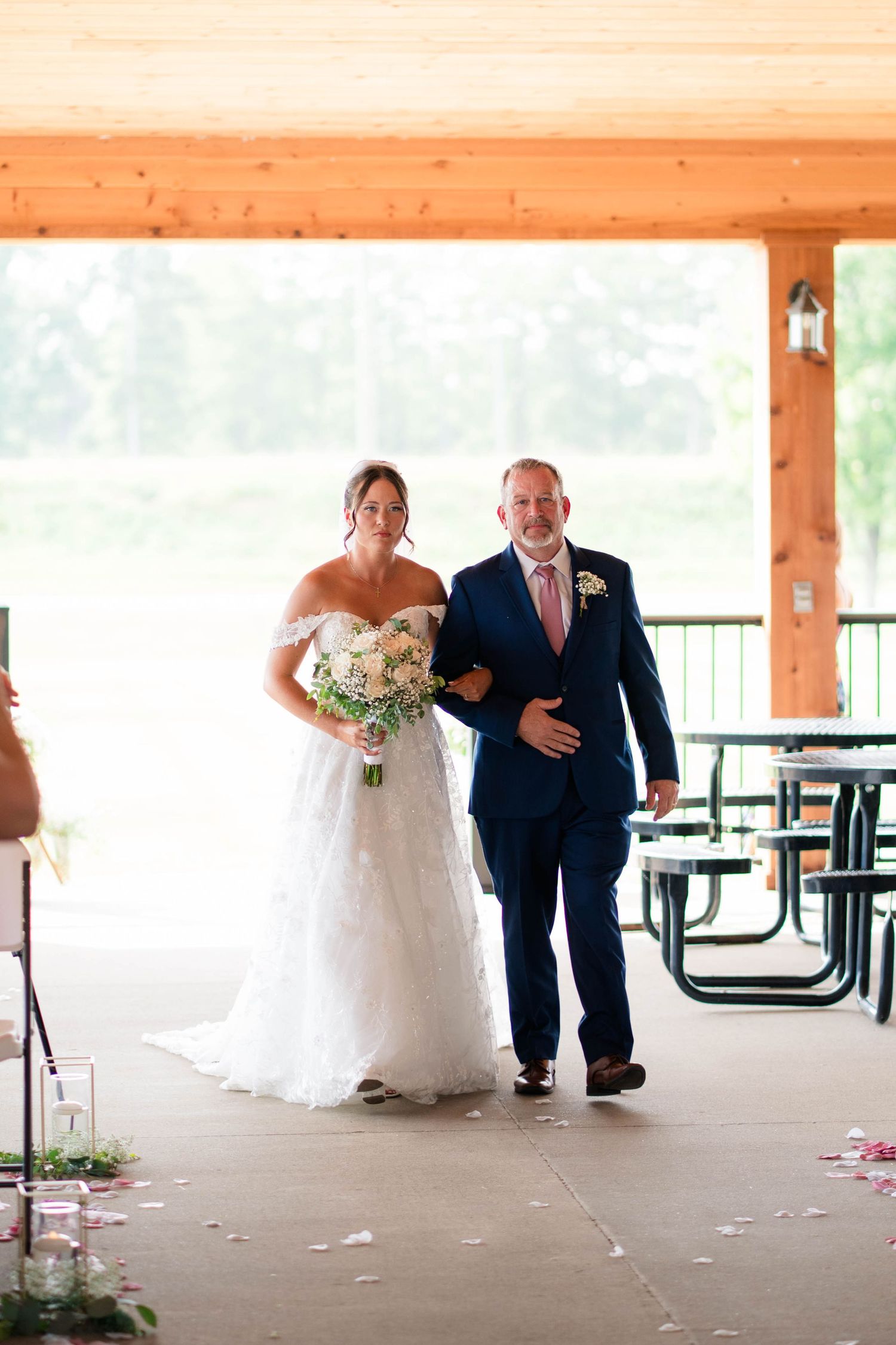 Bride in white off-shoulder gown holds bouquet while being escorted down aisle by man in navy suit.
