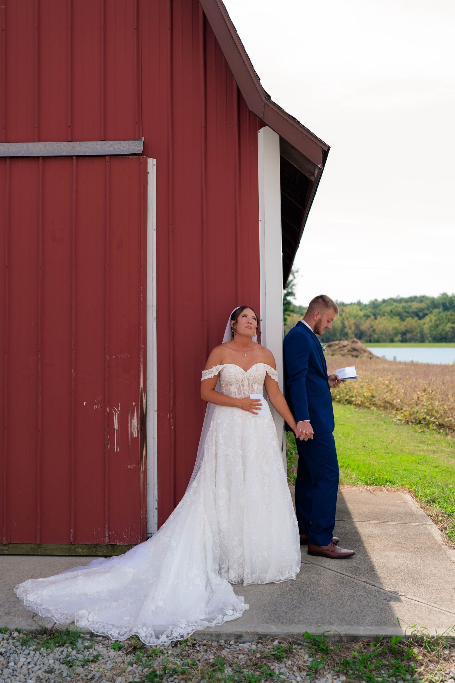 Bride and groom share a private first look moment beside a rustic red barn near a scenic waterfront.