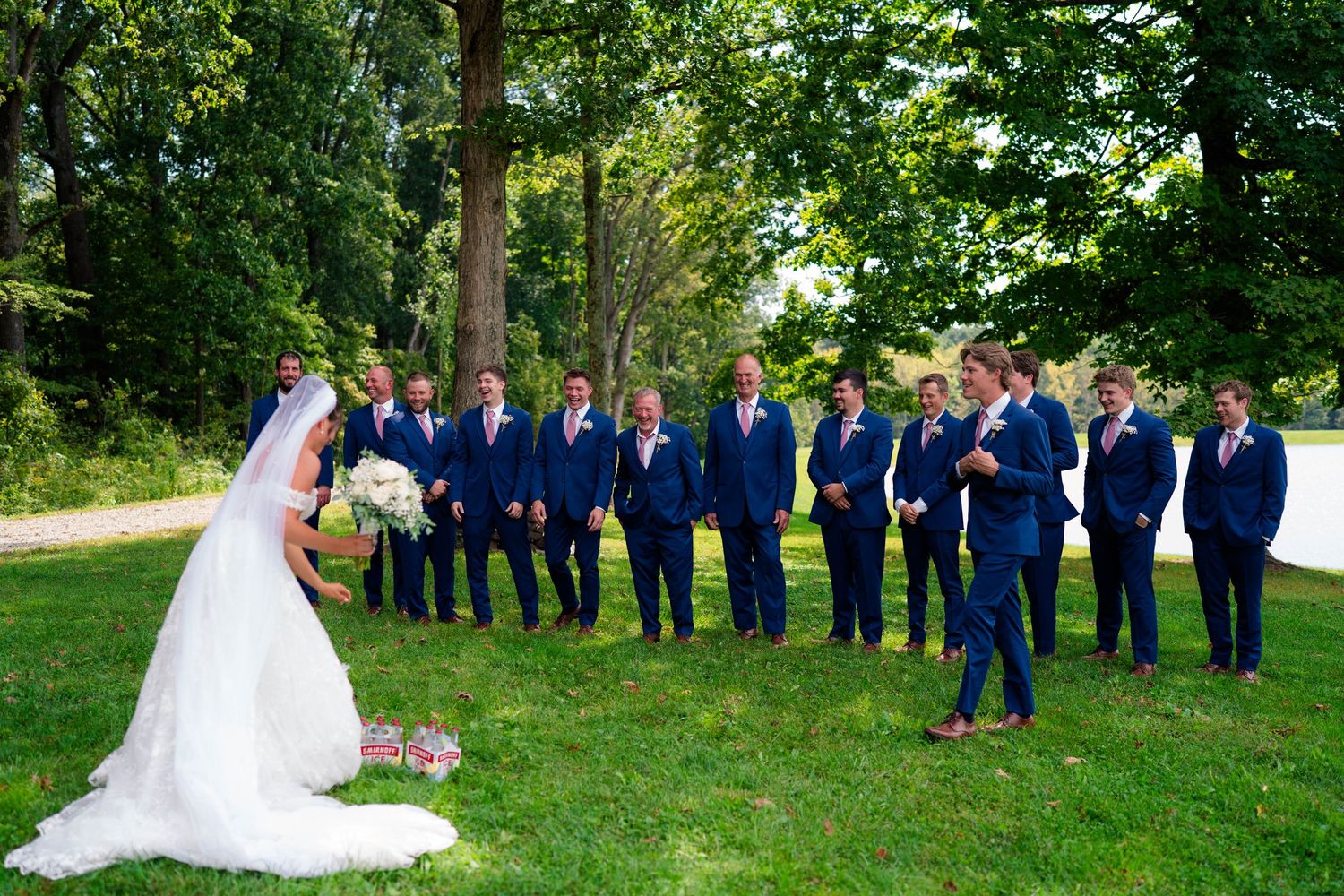 Bride approaches groomsmen in navy suits lined up on a green lawn surrounded by tall trees.