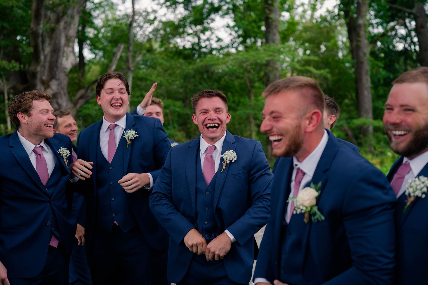 Groomsmen in navy blue suits with pink ties share a joyful laugh together outdoors at a wedding celebration.