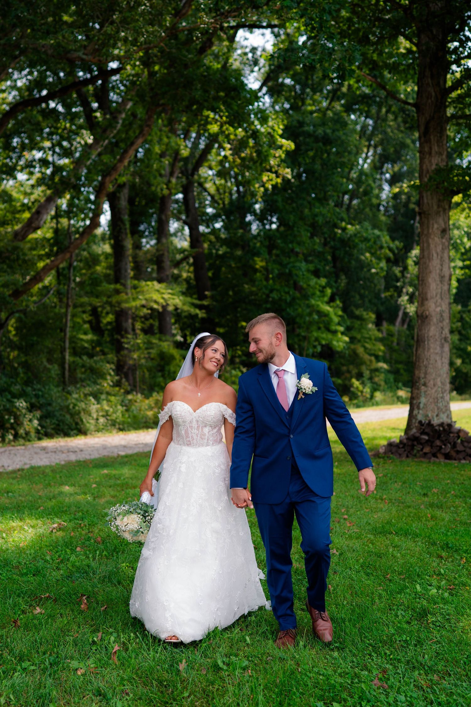 Bride and groom walk hand in hand through a lush green park, smiling at each other on their wedding day.