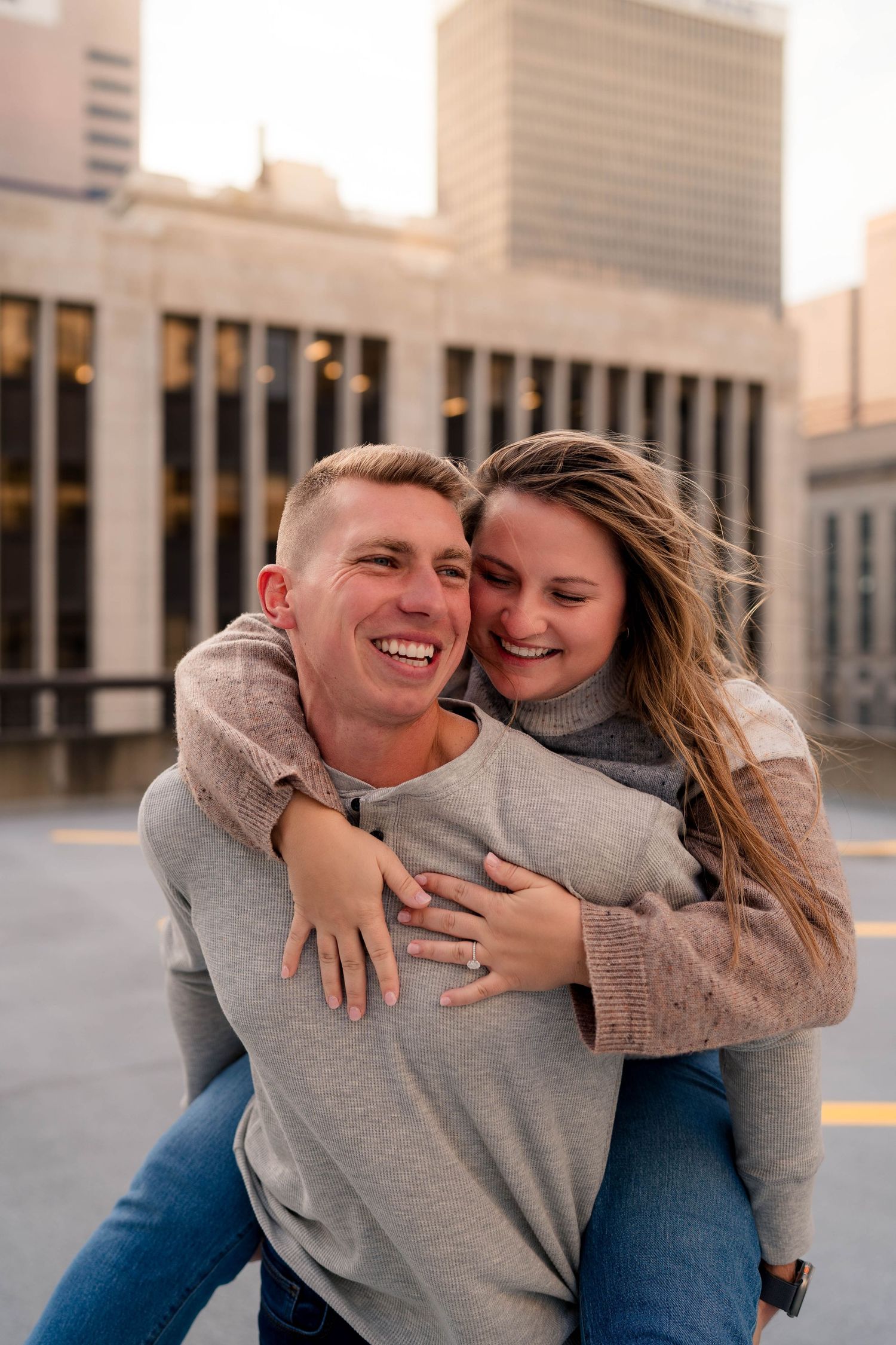 Joyful couple laughing as man gives woman a piggyback ride on a city rooftop at sunset.
