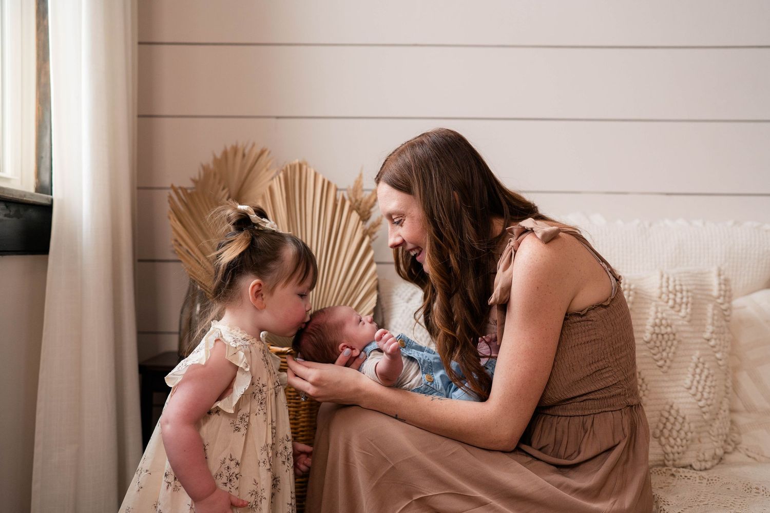 A mother smiles at her newborn baby while her young daughter looks on in a warm, bohemian-styled room.