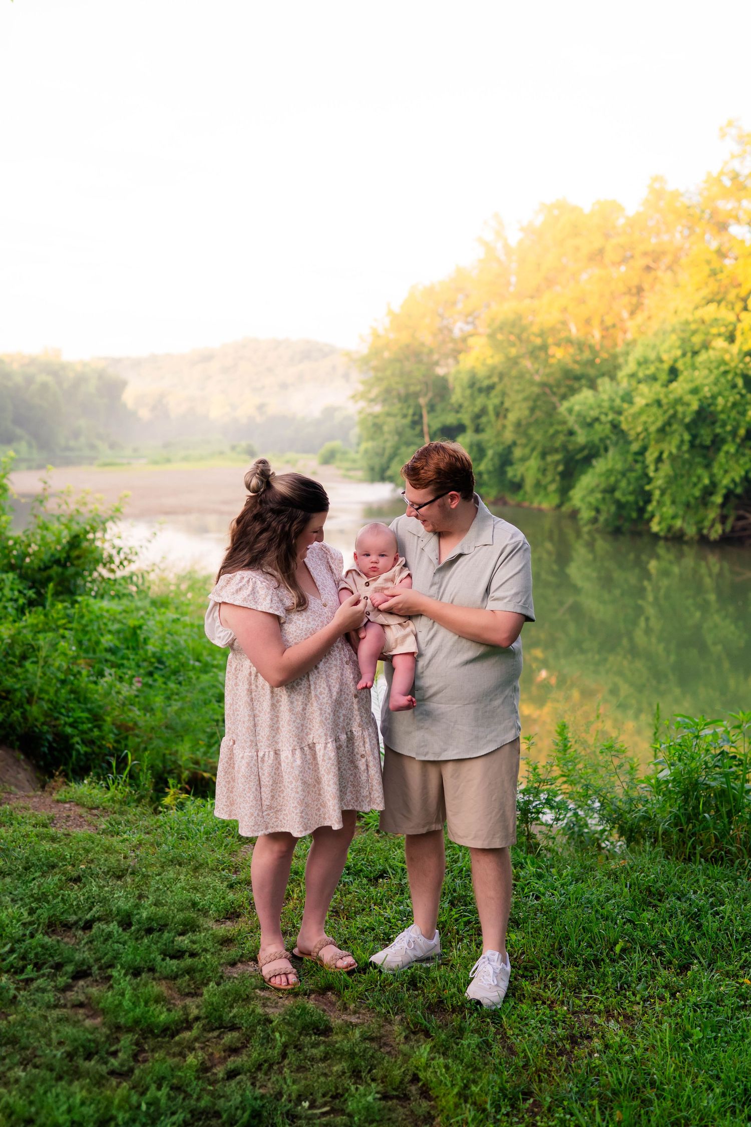 Young family of three standing by river at golden hour, parents admiring newborn baby.