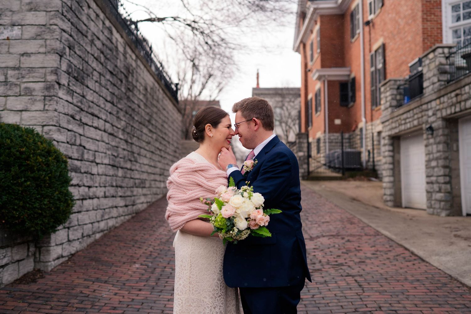 Bride and groom share a romantic moment on a brick alley, she holds a bouquet, wearing a pink shawl over her wedding dress.