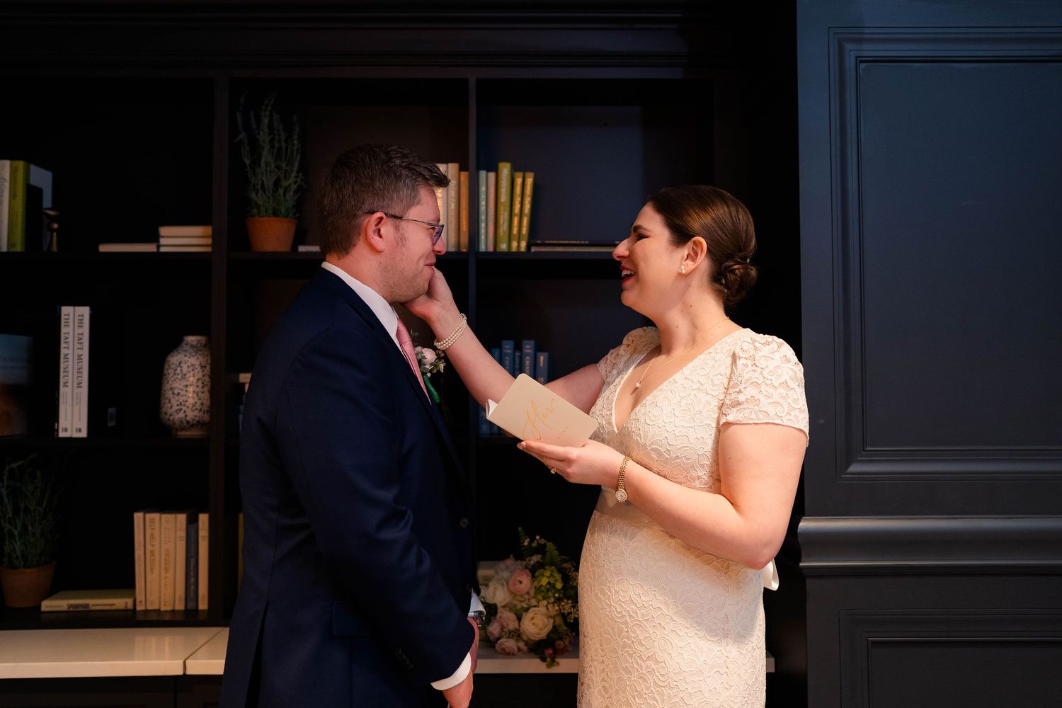 Emotional bride touching grooms face tenderly while reading wedding vows in dark elegant interior.