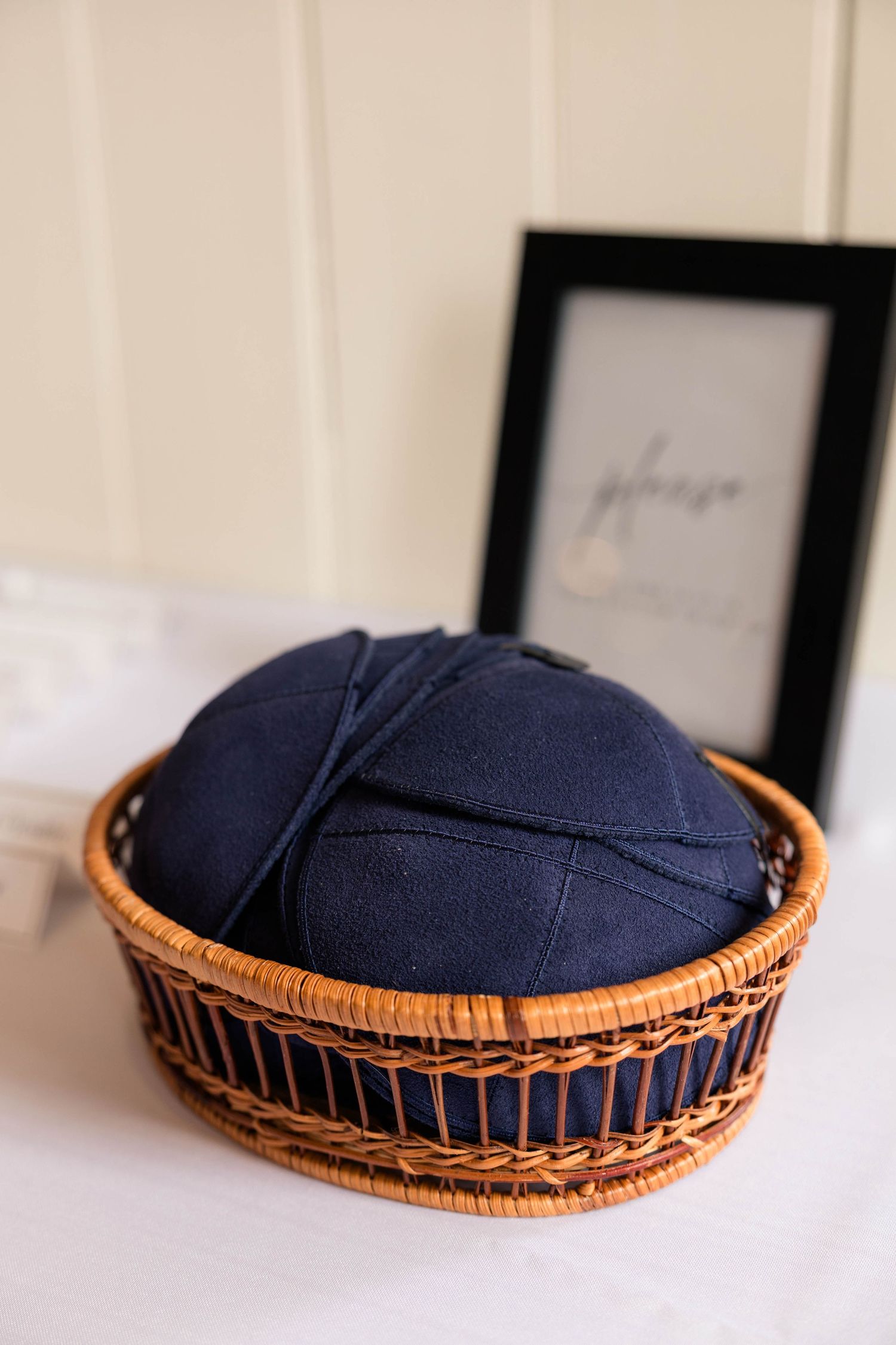 Wicker basket filled with navy blue kippot displayed on a white table with a framed sign at a Jewish wedding.