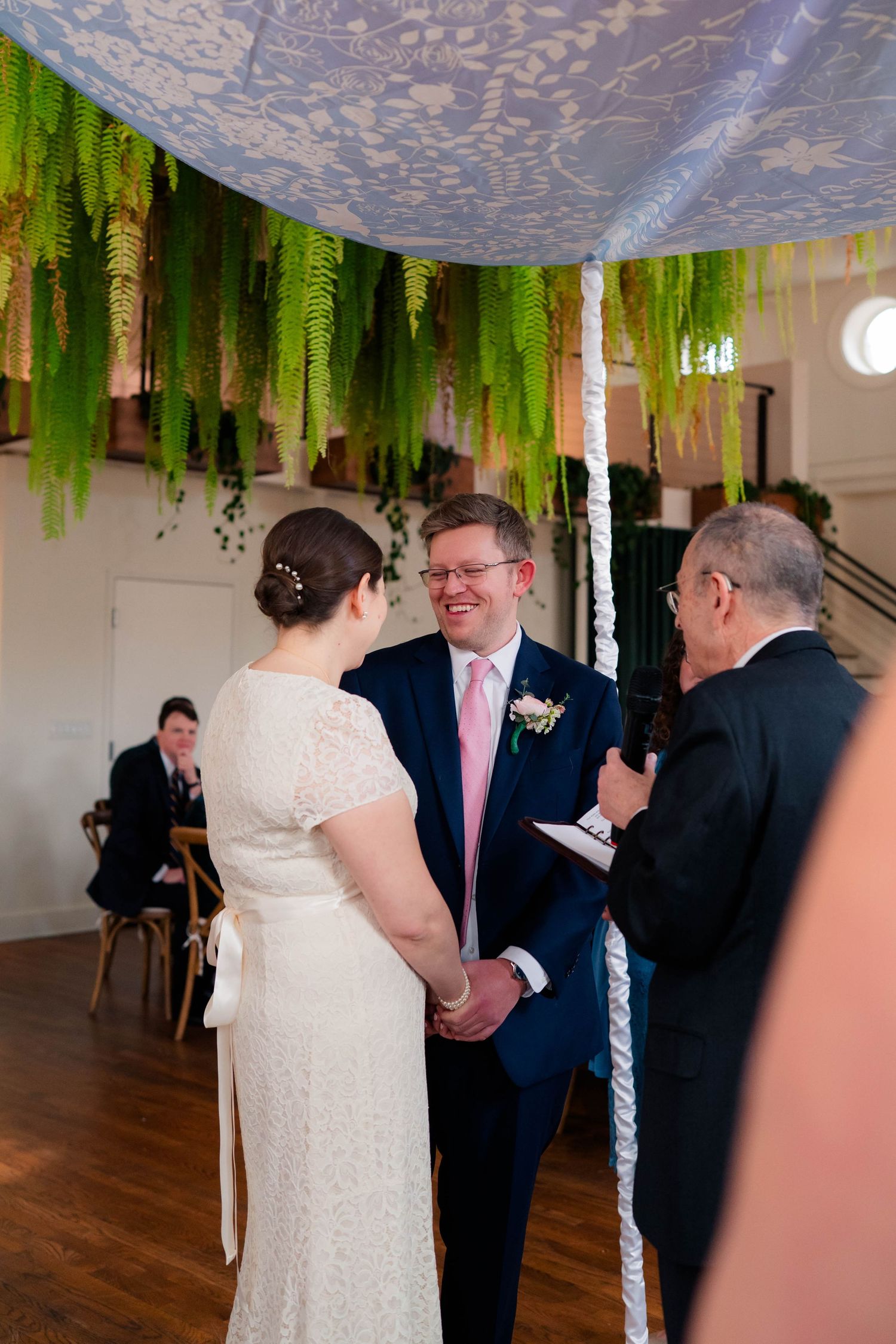 Bride and groom hold hands smiling during indoor Jewish wedding ceremony under floral chuppah with fern decorations.
