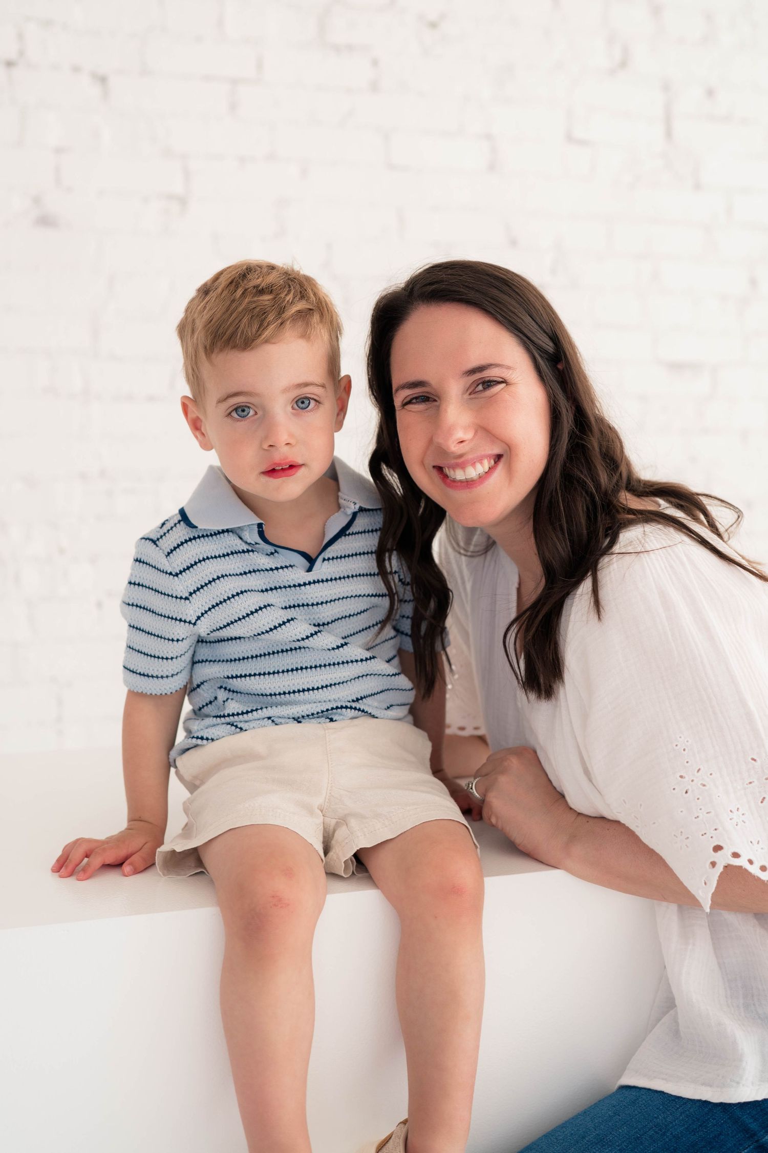 Smiling dark-haired mother posing with her young blonde son seated on a white bench against a brick wall.