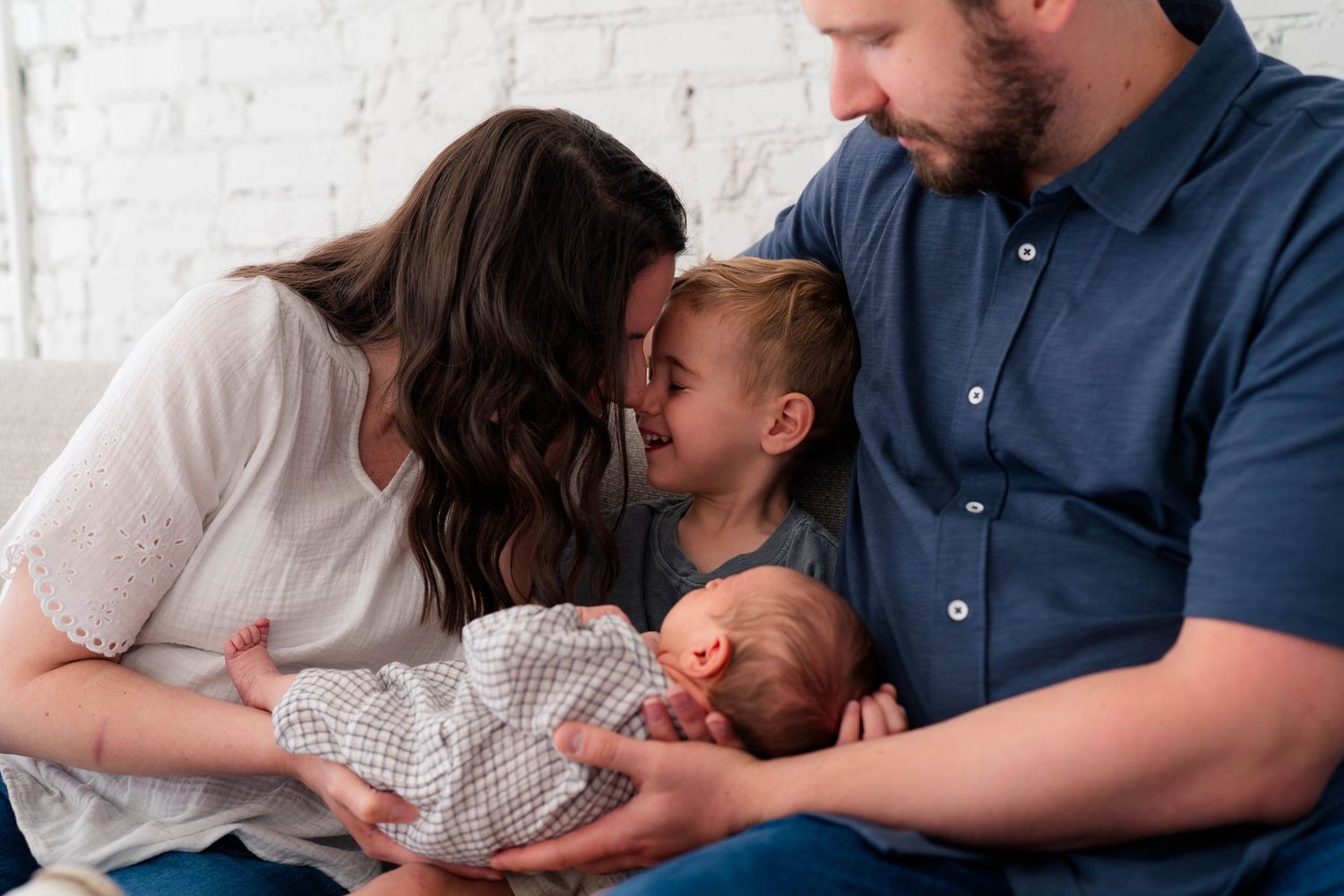 A loving family of four admires their newborn baby, with mom and toddler leaning in for a gentle kiss.
