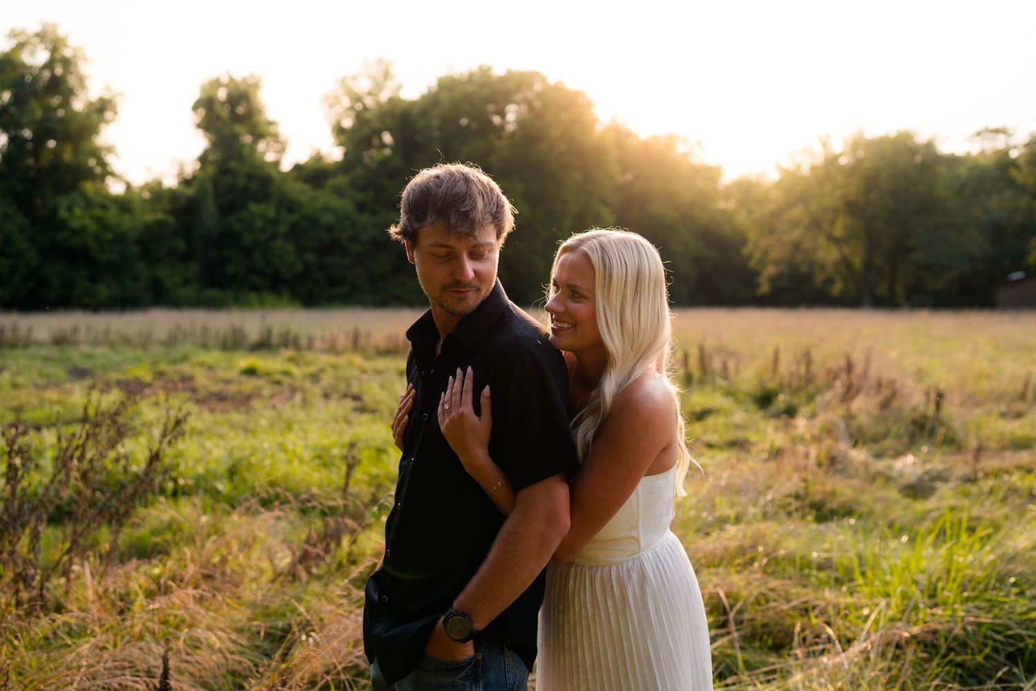 Couple embracing in golden sunset field, woman in white dress leaning against man in black shirt.