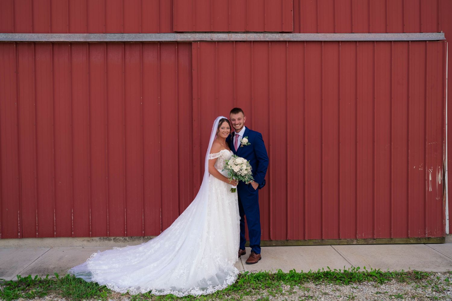 Newlyweds pose together smiling in front of a classic red barn wall on their wedding day.