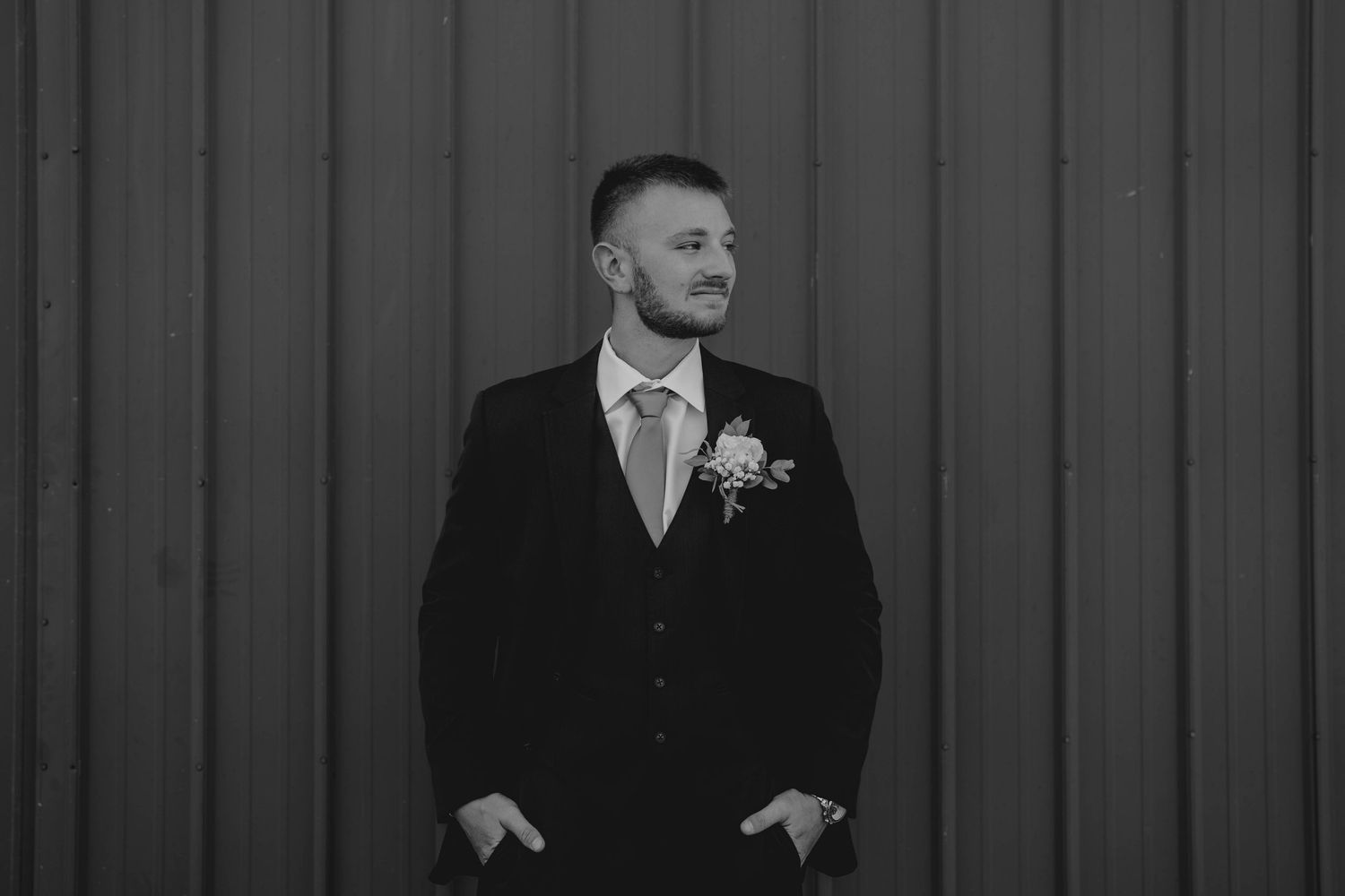 Groom in black suit with floral boutonniere posing against corrugated metal wall in black and white.