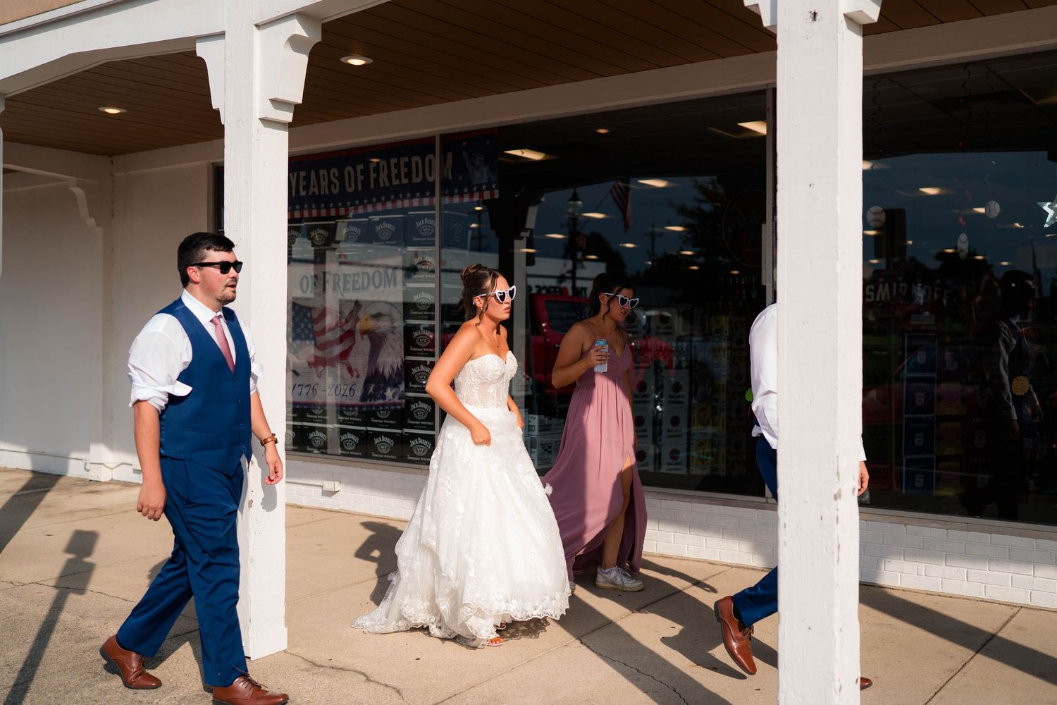 Bride in white gown walking with wedding party past storefront on sunny day.
