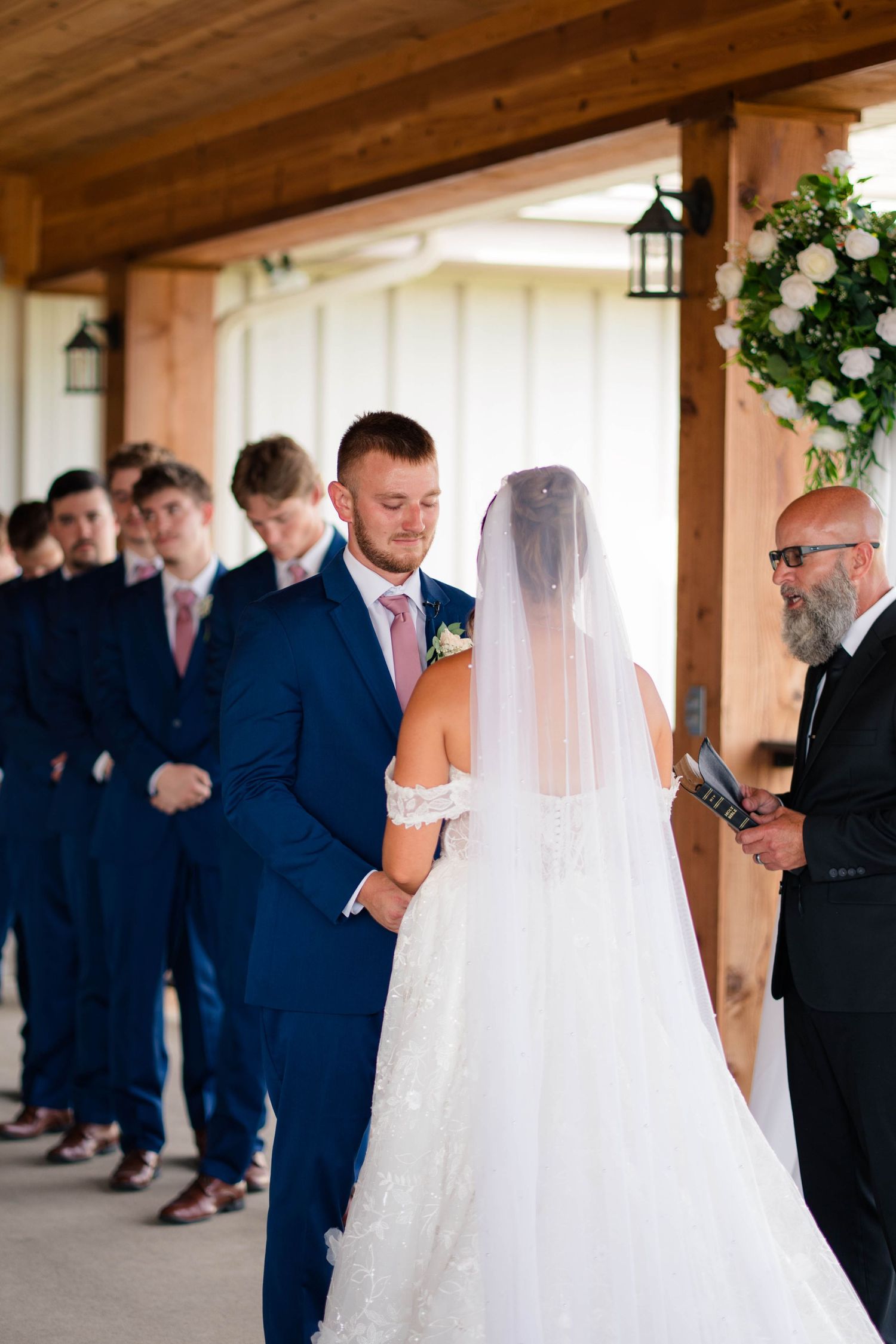 Groom holds brides hands during wedding ceremony with groomsmen lined behind.