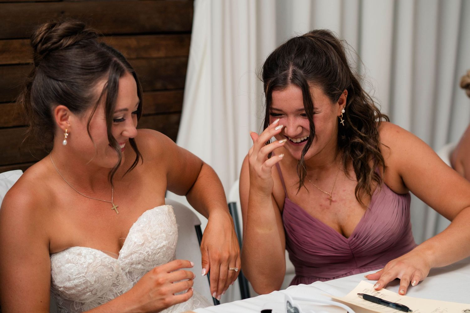 Bride and bridesmaid in purple dress laugh together while reading at wedding reception table.