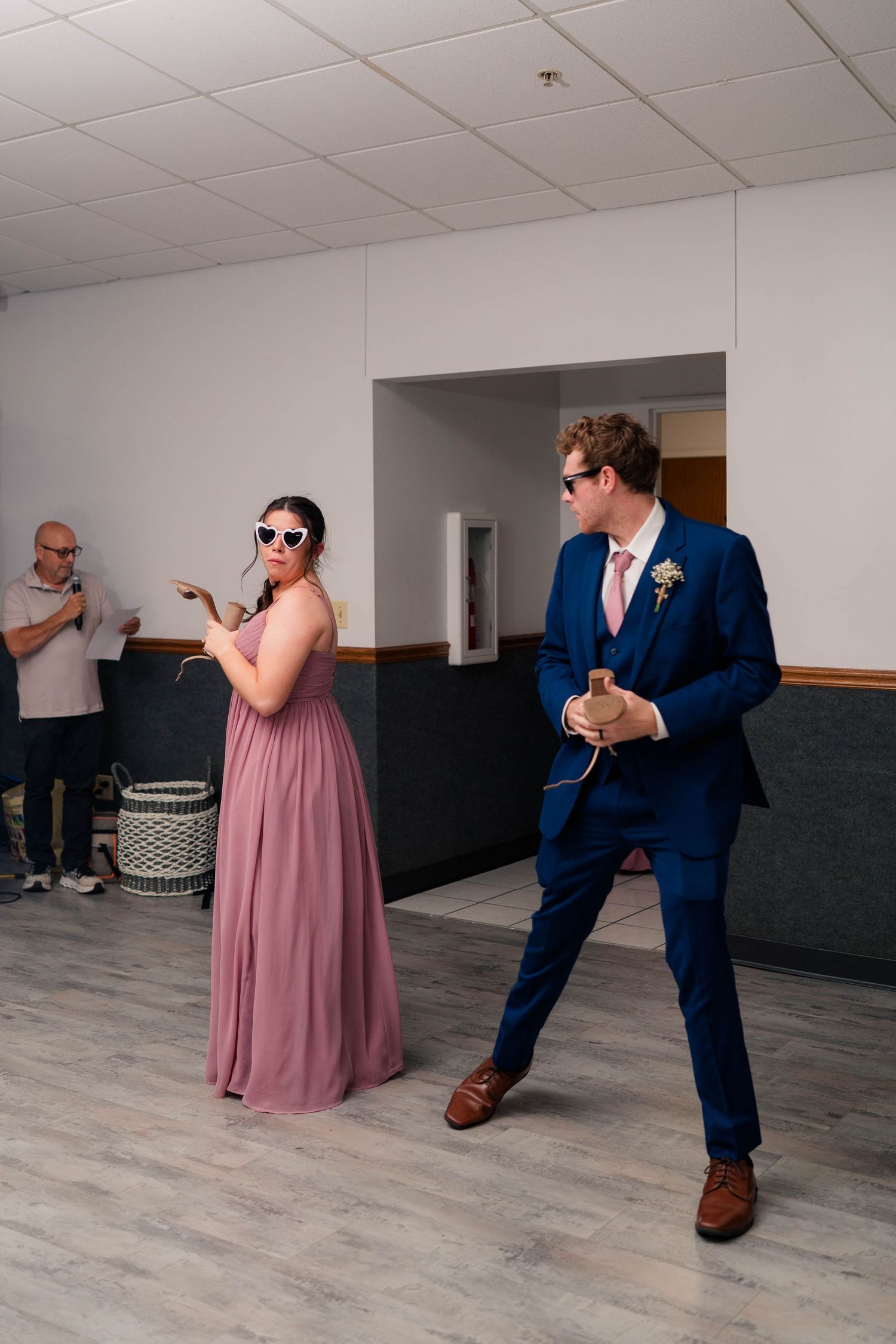 Bridesmaid in pink gown and groom in navy suit dance playfully at wedding reception indoor venue.