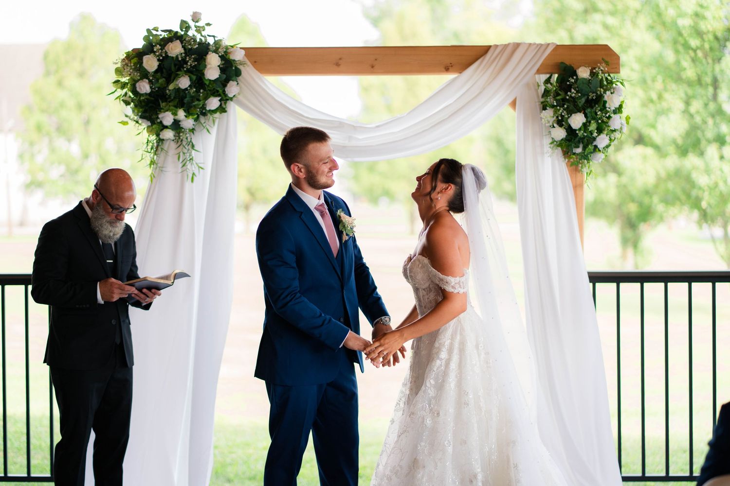 Bride and groom hold hands at floral arch during outdoor wedding ceremony.