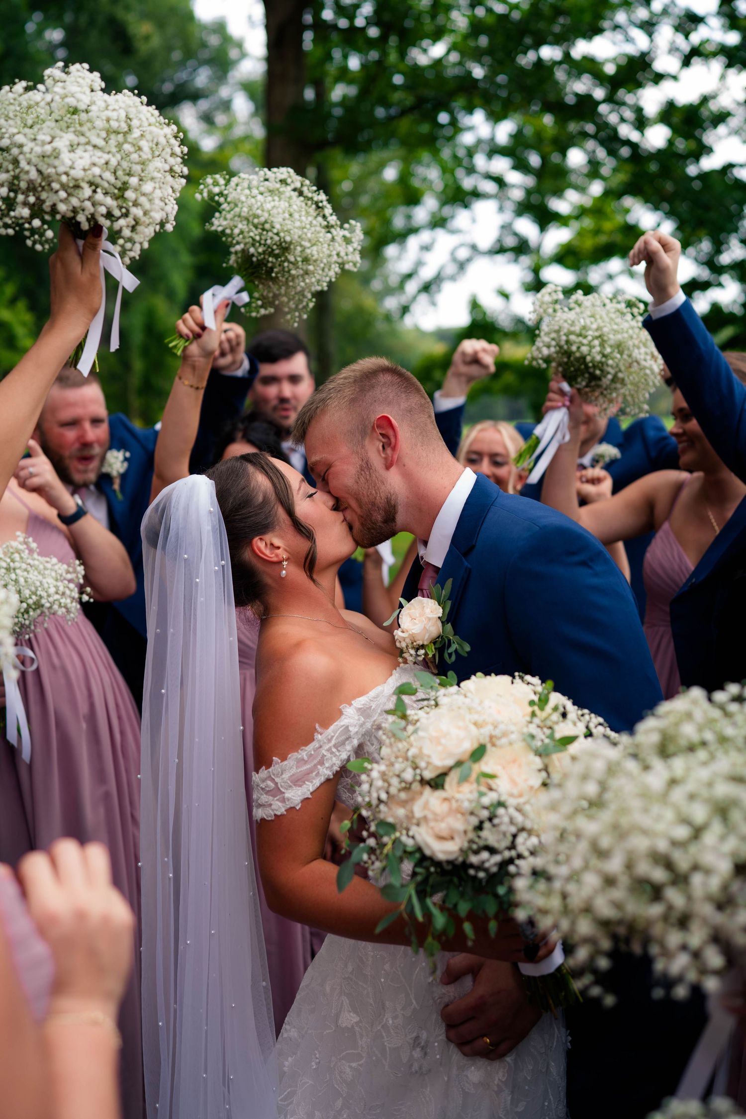 Bride and groom share a kiss surrounded by cheering wedding party holding baby's breath bouquets outdoors.