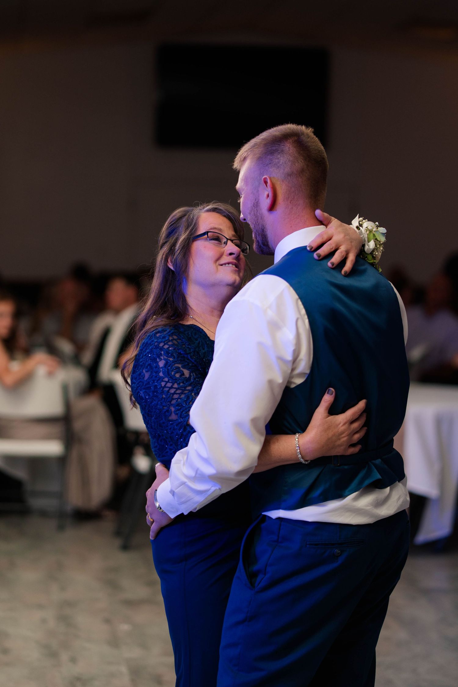 Woman in navy lace dress smiles warmly while dancing with groom in blue suit at wedding reception.