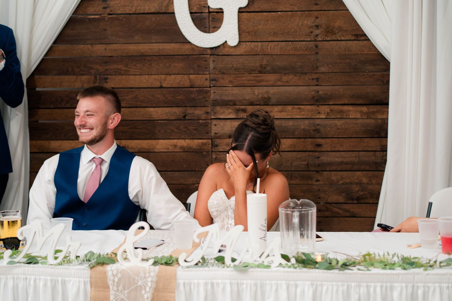 Bride laughing and covering face while groom smiles during wedding reception speech at rustic wood backdrop venue.