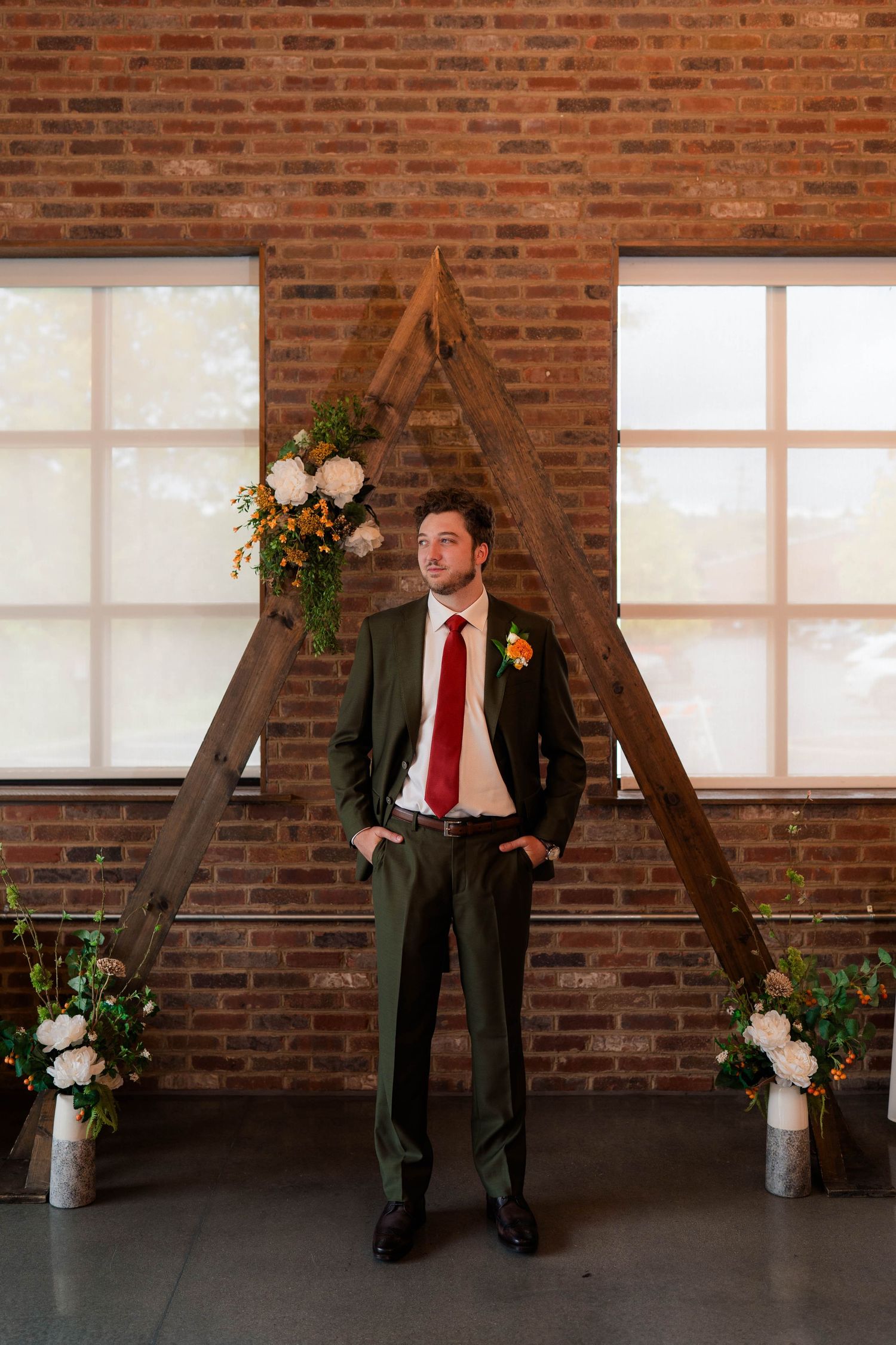 Groom in olive suit with red tie stands before wooden triangle arch at brick wall wedding venue.