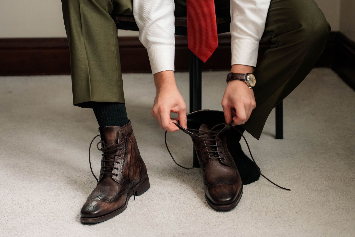 Man in dress pants and red tie tying dark brown leather lace-up boots while seated, wearing a watch.