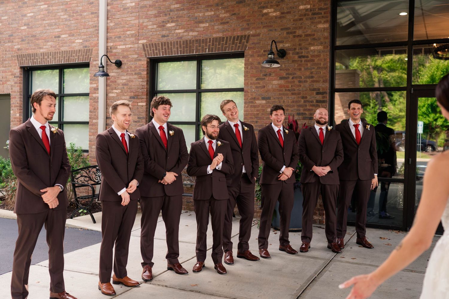 Groomsmen in dark brown suits with red ties stand outside brick building awaiting bride's reveal.