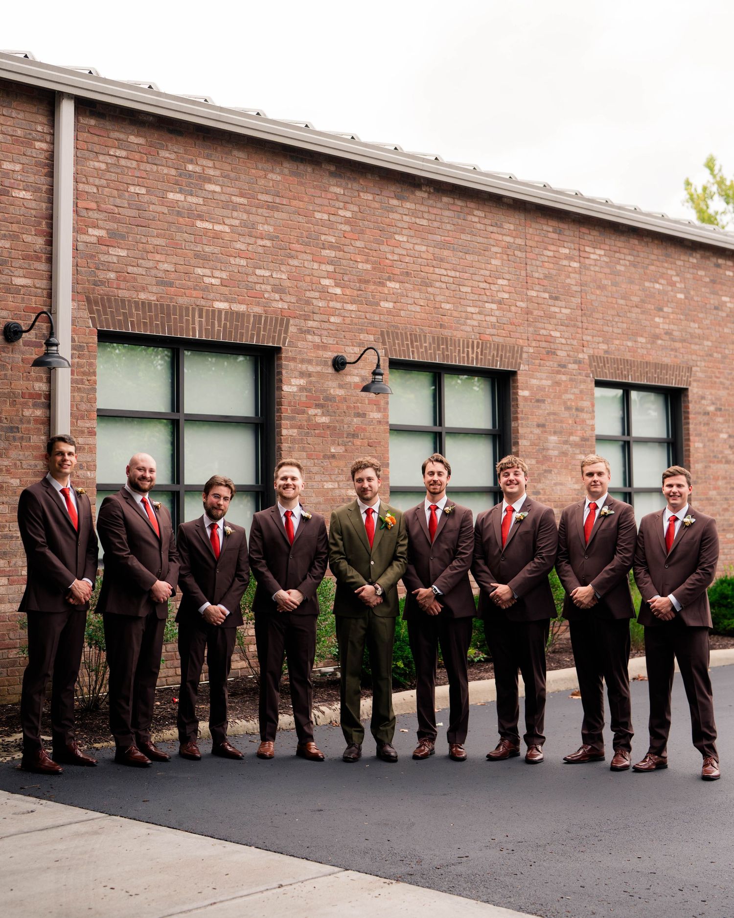 Groom and groomsmen in dark suits with red ties pose in a line outside brick building.