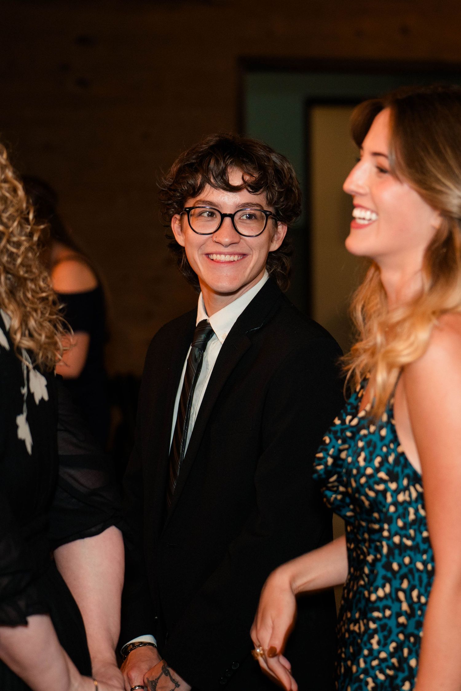Young man in black suit and glasses smiling at wedding reception with guests in background.