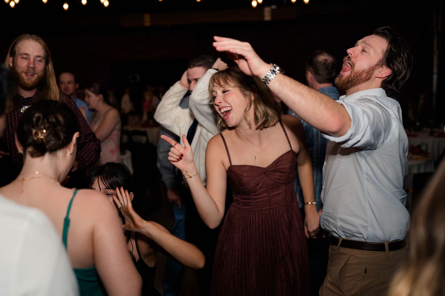 Guests laughing and dancing energetically at wedding reception under warm string lights in dark venue.