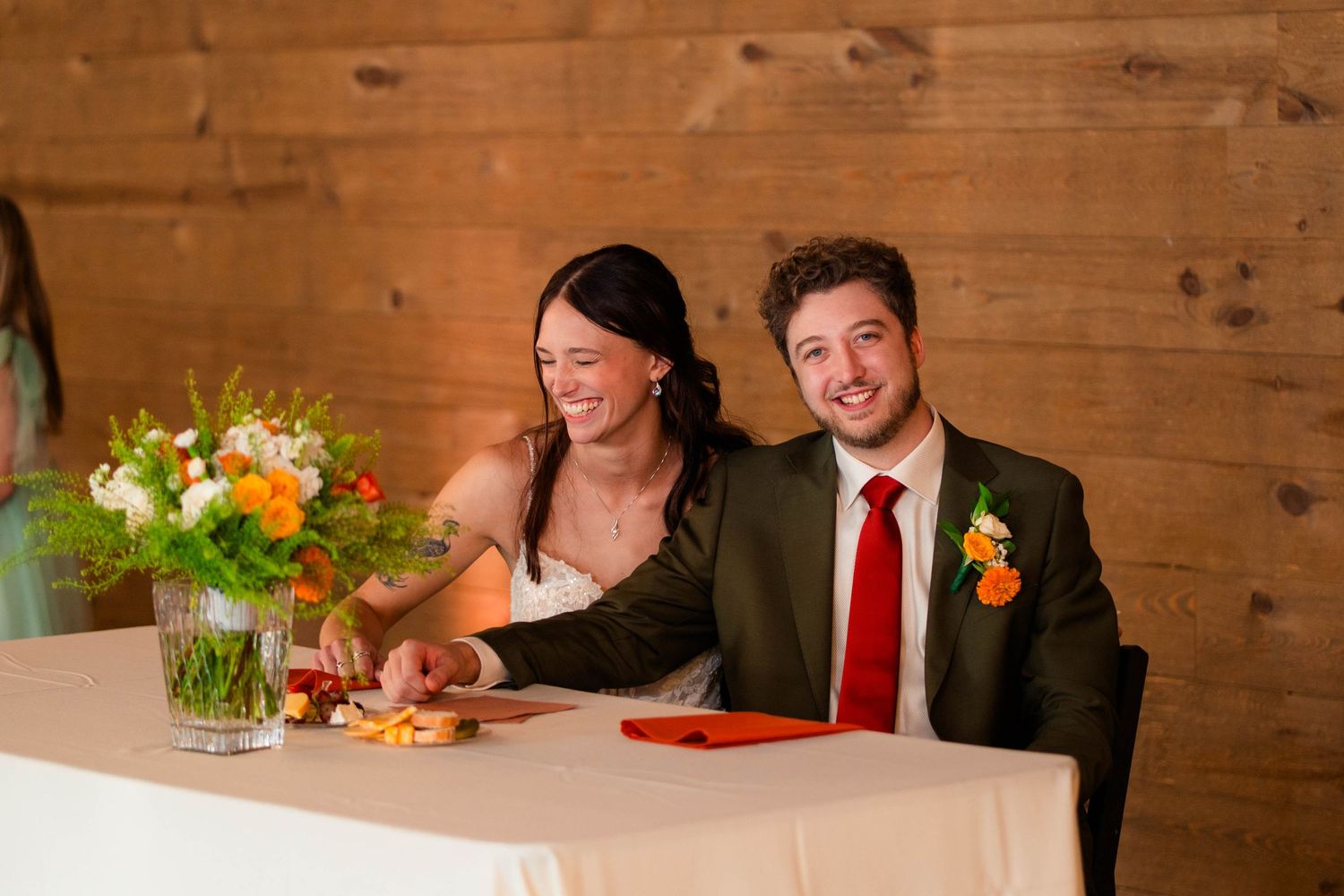 Bride and groom smile together at sweetheart table with orange and white floral arrangement.