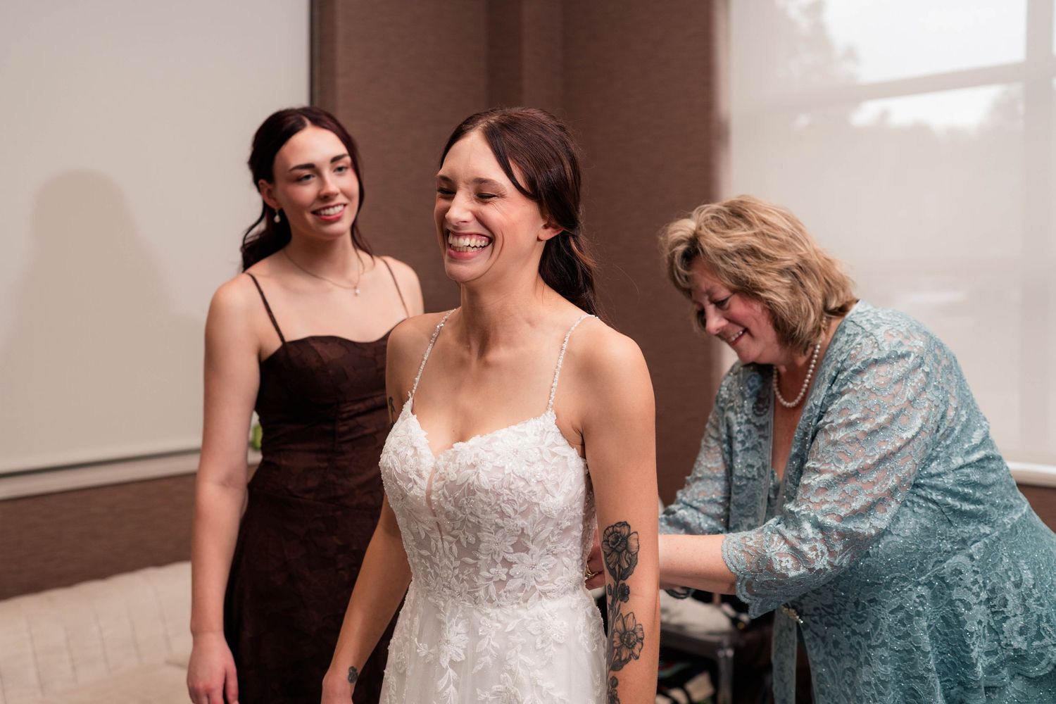 Bride smiles in white lace gown as mother helps with dress while bridesmaid looks on.