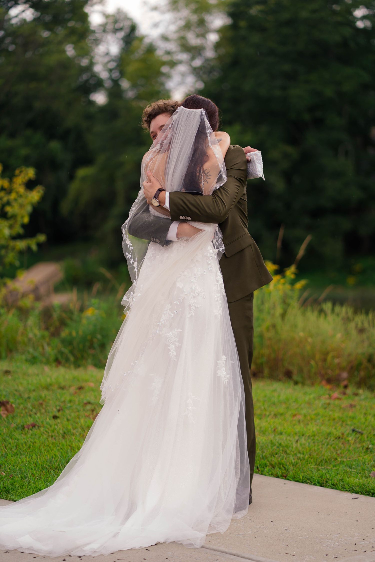 Bride and groom embracing and kissing behind veil in lush green outdoor setting.