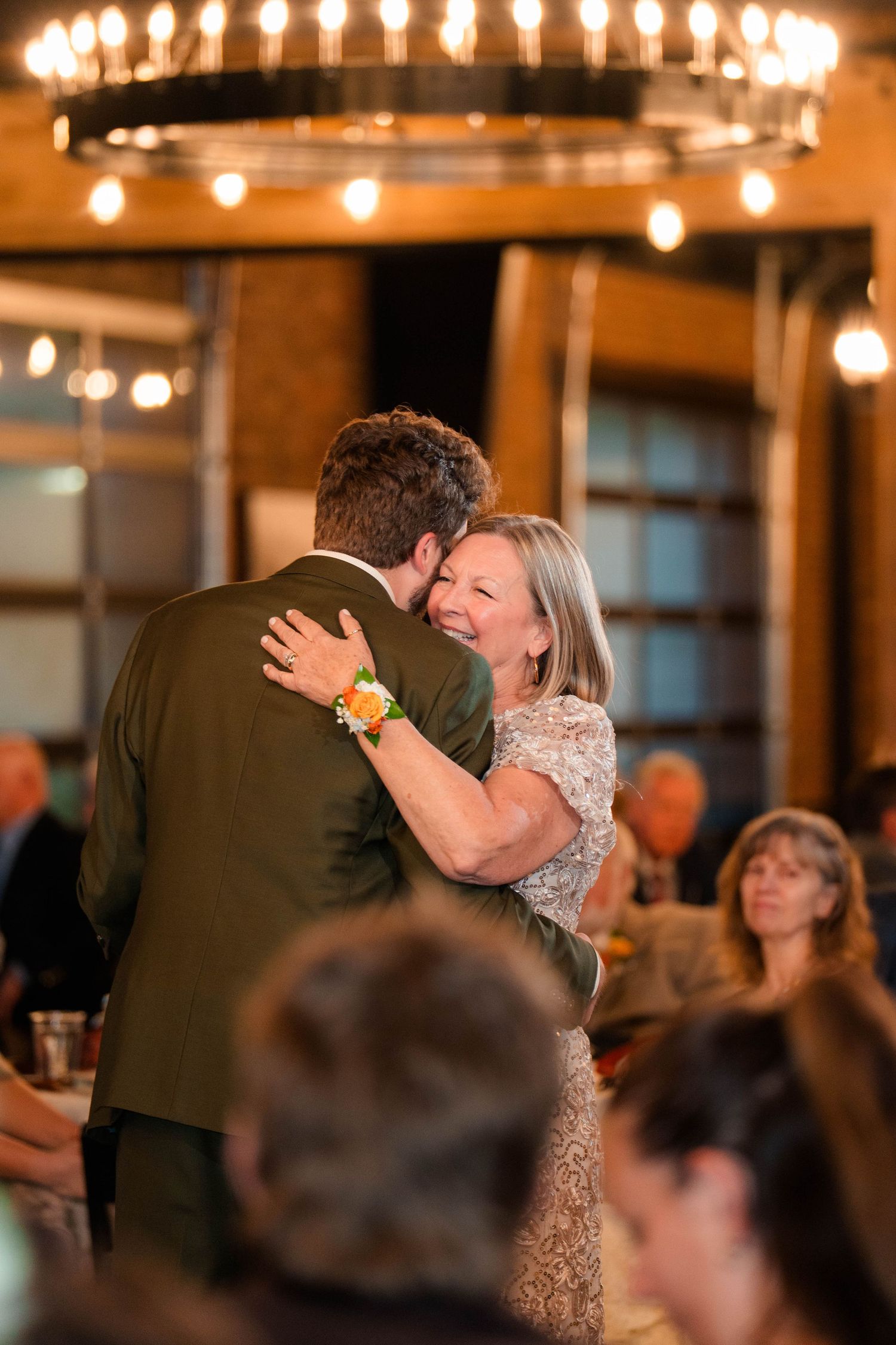Groom and mother share a joyful dance under chandelier at wedding reception.