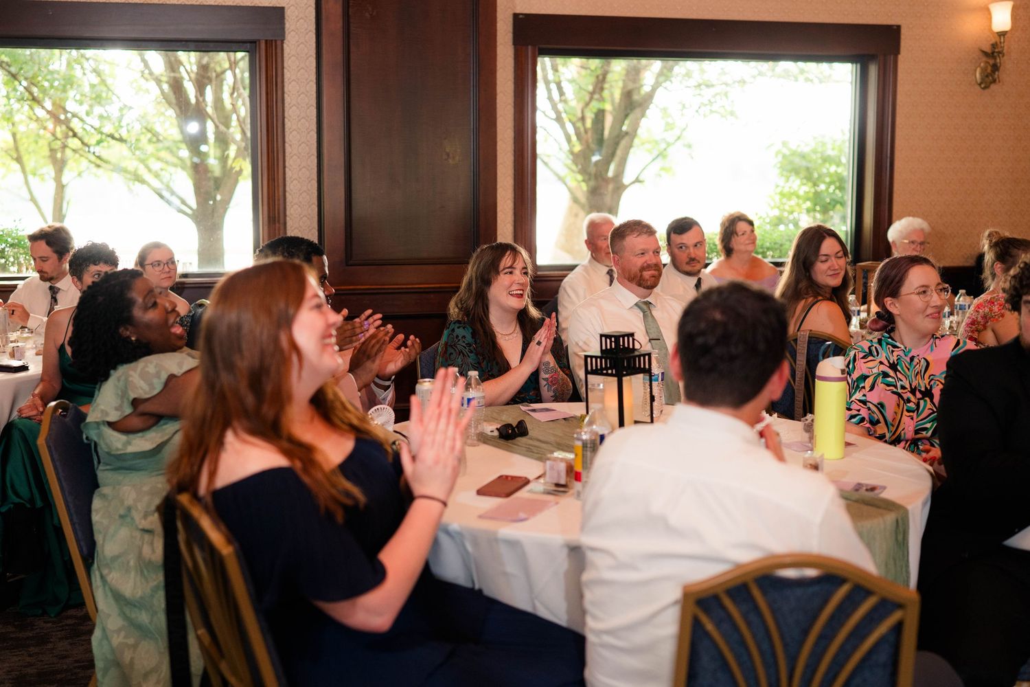 Wedding reception guests laugh and applaud at round tables in bright banquet hall.